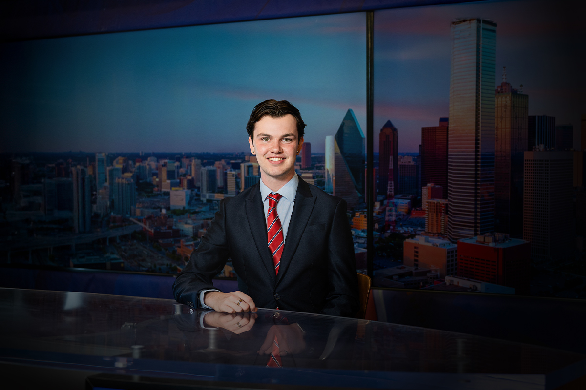 Man in a suit and tie sitting at a table with a cityscape background behind him